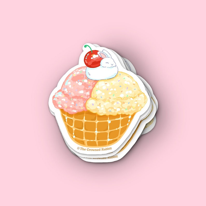 Peut inclure: Un autocollant repr&eacute;sentant un c&ocirc;ne de cr&egrave;me glac&eacute;e dessin&eacute; avec deux boules de cr&egrave;me glac&eacute;e, une rose et une jaune, surmont&eacute;es d'une cerise et d'un lapin blanc. Le c&ocirc;ne est de style gaufr&eacute; et la cr&egrave;me glac&eacute;e est d&eacute;cor&eacute;e de p&eacute;pites. L'autocollant porte le texte "@The Crowned Rabbit" en bas.
