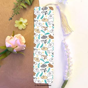 Op de afbeelding: Witte bladwijzer met een herhalend patroon van schattige cartoon konijnen in verschillende kleuren, waaronder bruin, grijs en wit. De konijnen zijn omringd door groene bladeren en gele bloemen. De bladwijzer heeft een witte kwast en de tekst "The Crowned Rabbit" onderaan.