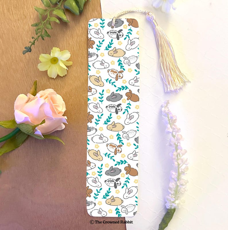 Op de afbeelding: Witte bladwijzer met een herhalend patroon van schattige cartoon konijnen in verschillende kleuren, waaronder bruin, grijs en wit. De konijnen zijn omringd door groene bladeren en gele bloemen. De bladwijzer heeft een witte kwast en de tekst "The Crowned Rabbit" onderaan.