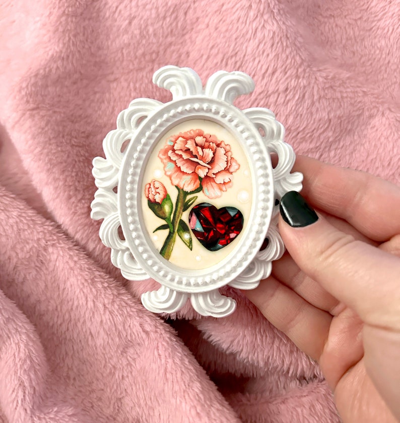 May include: A white ornate frame with a pink carnation and a red heart-shaped gem inside. The frame is held in a hand.