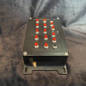 May include: A black box with 16 red buttons and one black button. The box has a metal bracket on the bottom.