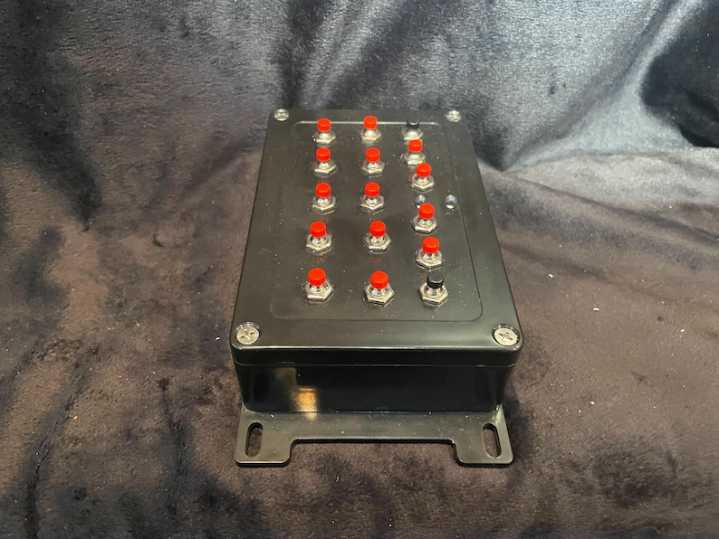 May include: A black box with 16 red buttons and one black button. The box has a metal bracket on the bottom.