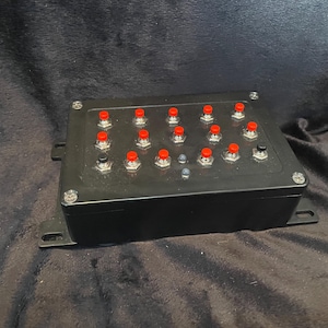 May include: A black box with 18 red buttons and a single black button. The buttons are arranged in a grid pattern.