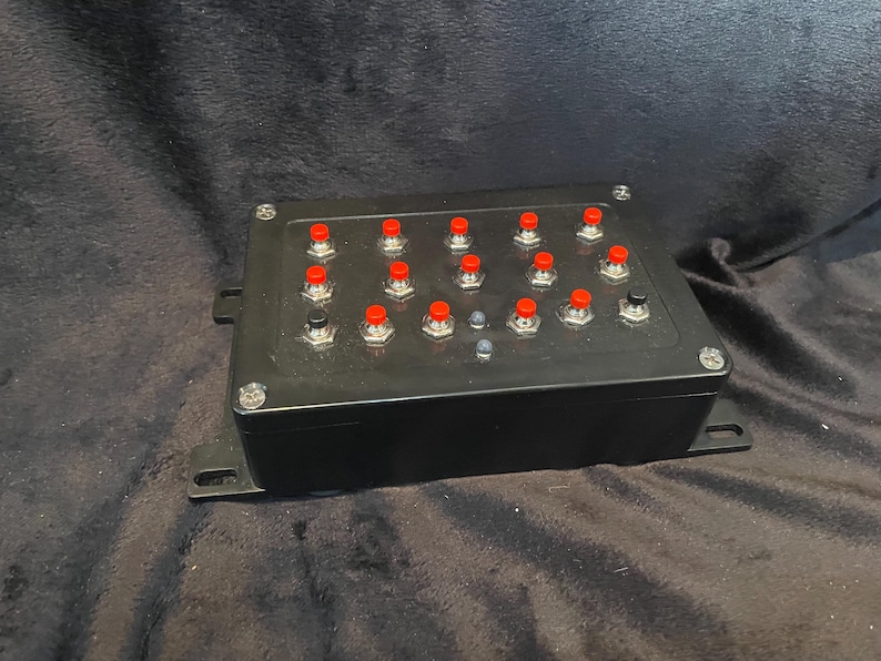 May include: A black box with 18 red buttons and a single black button. The buttons are arranged in a grid pattern.