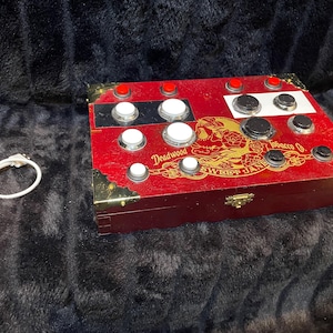 May include: A red wooden box with various buttons and a gold design that reads "Deadwood Sweet Jane Tobacco Co." The box has gold corner protectors and a small clasp. A white cable is next to the box.