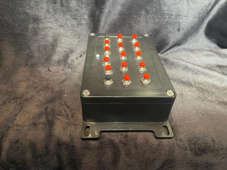 May include: A black box with 16 red buttons. The box has a metal bracket on each side.