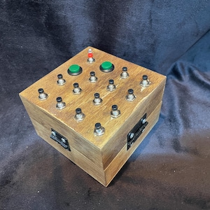 May include: A wooden box with a hinged lid, featuring a grid of 16 black push buttons and two green push buttons. The box has a red LED light on the top.