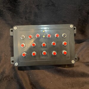 May include: Black control panel with 16 red push buttons and two black buttons. The panel has a black plastic casing with a screw on each side.