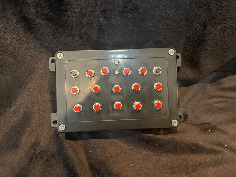May include: Black control panel with 16 red push buttons and two black buttons. The panel has a black plastic casing with a screw on each side.