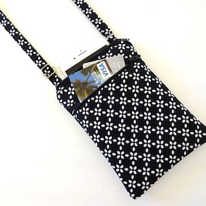 Op de afbeelding: Een zwart-witte crossbody telefoontas met bloemenpatroon en ritssluiting. De tas heeft een zwarte band en een goudkleurige gesp. Een witte smartphone en creditcards zijn zichtbaar in de tas.