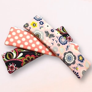 May include: Four fabric eyeglass cases in various patterns. One is coral with white polka dots, another is black with a floral design, and two are pink with floral and butterfly patterns. The cases are rectangular with a triangular shape at the end.