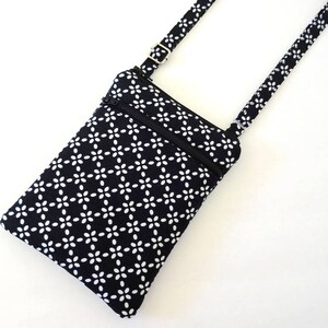Op de afbeelding: Een zwart-witte crossbody tas met een bloemenpatroon. De tas heeft een rechthoekige vorm met een ritssluiting aan de bovenkant en een verstelbare riem. De riem en de stof van de tas hebben een herhalend wit bloemmotief op een zwarte achtergrond.