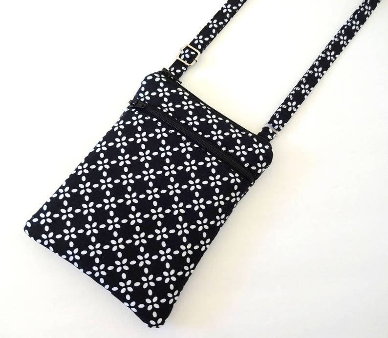 Op de afbeelding: Een zwart-witte crossbody tas met een bloemenpatroon. De tas heeft een rechthoekige vorm met een ritssluiting aan de bovenkant en een verstelbare riem. De riem en de stof van de tas hebben een herhalend wit bloemmotief op een zwarte achtergrond.