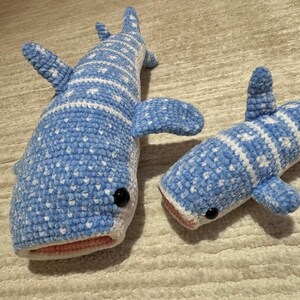 May include: Two handmade crochet whale shark plushies. The plushies are blue with white markings and black eyes. The larger plushie is approximately 24 inches long, and the smaller one is about 16 inches long. The plushies have a pink mouth.