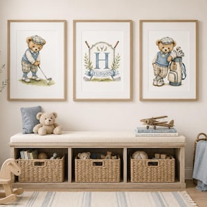 Op de afbeelding: Drie ingelijste wandplaten met aquarelillustraties van een teddybeer die golf speelt, een centraal wapenschild met de letter H en de naam HUDSON, en een teddybeer die tegen een golftas leunt, boven een houten opbergbank geplaatst.