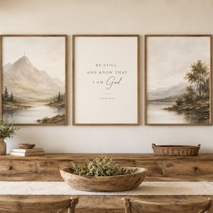 May include: A set of three framed wall art prints featuring two landscape paintings of mountains and a lake flanking a central print with the text BE STILL, AND KNOW THAT I AM God. PSALM 46:10 in a neutral color palette with wooden frames.
