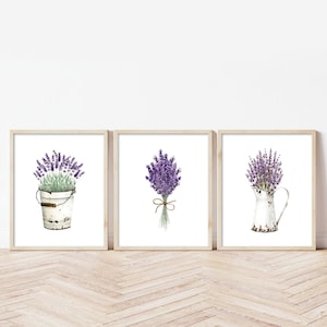 Op de afbeelding: Set van drie ingelijste prints met aquarel illustraties van lavendelbloemen in verschillende arrangementen. De eerste print toont een boeket lavendel in een witte metalen emmer. De tweede print toont een boeket lavendel gebonden met touw. De derde print toont een boeket lavendel in een witte kan.