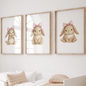 May include: Three framed prints featuring watercolor illustrations of bunnies with pink bows. The bunnies are light brown with long ears and are set against a white background. The frames are light wood, and the prints are displayed above a white bed with beige bedding.