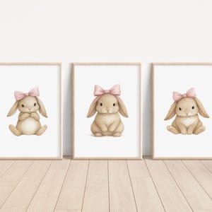 May include: Three framed prints featuring watercolor illustrations of bunnies. Each bunny is light brown with a pink bow on its head. The bunnies are in different poses, sitting or crouching. The frames are light wood, and the background is white.