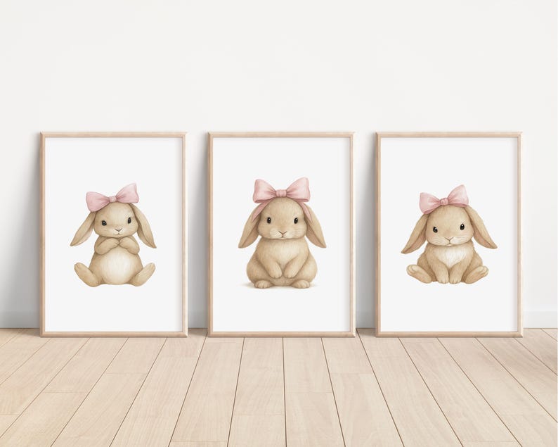 May include: Three framed prints featuring watercolor illustrations of bunnies. Each bunny is light brown with a pink bow on its head. The bunnies are in different poses, sitting or crouching. The frames are light wood, and the background is white.