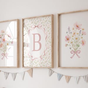 May include: Three framed floral art prints. Two prints feature bouquets of pink and white flowers tied with pink ribbons. The center print has a pink letter "B" inside a pink ribbon-edged oval. A banner of small flags hangs below.