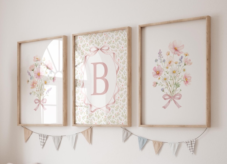 May include: Three framed floral art prints. Two prints feature bouquets of pink and white flowers tied with pink ribbons. The center print has a pink letter "B" inside a pink ribbon-edged oval. A banner of small flags hangs below.