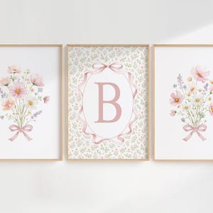 May include: Three framed floral art prints. Two prints feature watercolor bouquets of pink, white, and lavender flowers tied with pink ribbons. The center print has a pink letter "B" inside a pink ribbon frame.