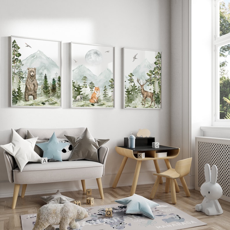 Op de afbeelding: Drie ingelijste aquarel illustraties van een beer, een vos en een hert in een bosrijke omgeving met bergen en volle maan. De illustraties hangen aan een witte muur in een kinderkamer met een grijze bank, een houten bureau en een stoel.
