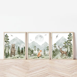 Op de afbeelding: Een set van drie ingelijste aquarelprints met een beer, vos en hert in een bosrijke omgeving met bergen en volle maan. De prints zijn in een licht houten lijst.