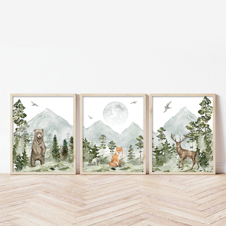 Op de afbeelding: Een set van drie ingelijste aquarelprints met een beer, vos en hert in een bosrijke omgeving met bergen en volle maan. De prints zijn in een licht houten lijst.