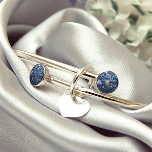 May include: A silver bracelet featuring two blue speckled stone details and a small heart charm. The bracelet is presented on a bed of shiny, silver fabric, with white flowers in the background.