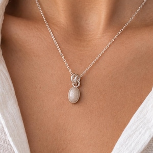 May include: A silver chain necklace featuring a small, oval-shaped pendant with a smooth, light-colored stone. The pendant is attached to the chain by a series of interconnected silver rings, creating a simple and clean design for the jewelry piece.