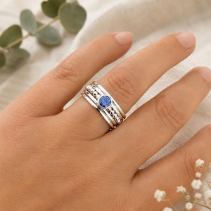 May include: A stackable ring set consisting of four individual silver bands. One band features a round, bezel-set blue gemstone, while another band has a textured beaded design. The remaining two bands are smooth, polished silver rings.