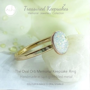 May include: A gold-colored ring featuring an oval-shaped stone with an opal sparkle finish. The text on the image reads: Treasured Keepsakes Memorial Jewellery Collection, The Oval Orb Memorial Keepsake Ring, Handmade in 14ct gold filled metal.