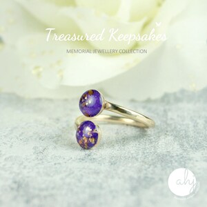 May include: A bypass ring featuring a metallic band and two circular purple cabochons with gold-colored flecks. The text "Treasured Keepsakes MEMORIAL JEWELLERY COLLECTION" is visible at the top, and a circular logo with "ahj" is at the bottom.