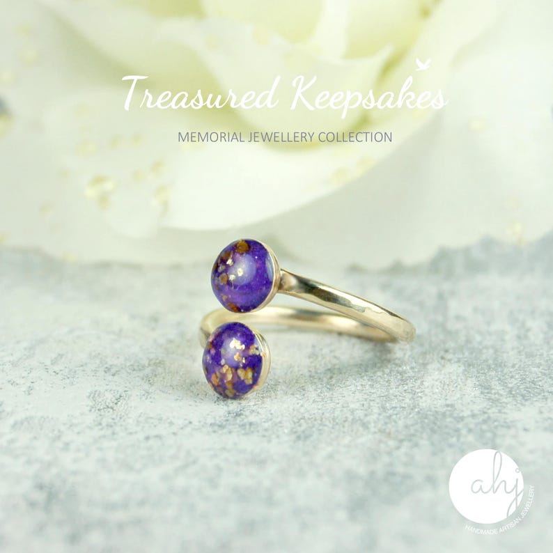 May include: A bypass ring featuring a metallic band and two circular purple cabochons with gold-colored flecks. The text "Treasured Keepsakes MEMORIAL JEWELLERY COLLECTION" is visible at the top, and a circular logo with "ahj" is at the bottom.