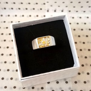 May include: A silver-toned ring with a rectangular gold-colored center filled with gold flakes. The ring is presented in a white box with a black interior, set against a polka-dot background. The ring's design is modern and eye-catching.