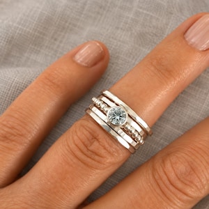 May include: A stack of four silver rings worn on a finger. The set includes two smooth bands, one band with a beaded texture, and one band featuring a central round, light blue gemstone set in a bezel. The metal has a polished, reflective finish.