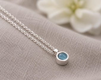 May include: A silver necklace featuring a small, circular pendant with a bezel-set, light blue stone. The pendant is attached to a delicate silver chain with round links. The metal has a polished, reflective finish, and the stone has a smooth, slightly textured surface.