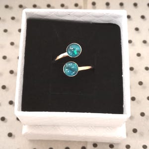 May include: A bypass ring featuring two circular settings with a dark teal, iridescent, glitter-like inlay. The ring has a polished gold-tone metal band and is presented inside a square white box with a black velvet insert.