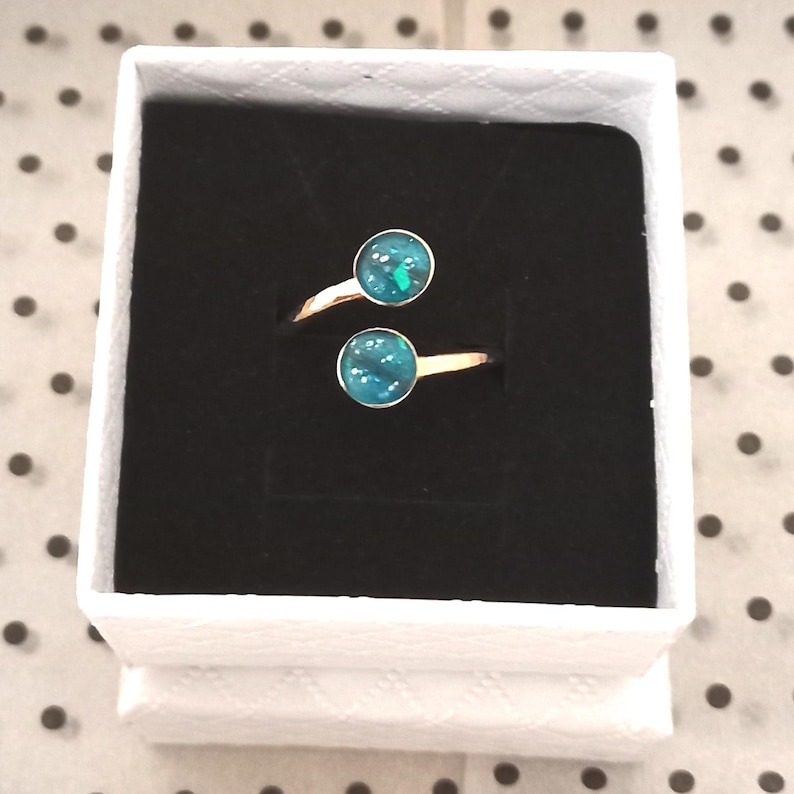 May include: A bypass ring featuring two circular settings with a dark teal, iridescent, glitter-like inlay. The ring has a polished gold-tone metal band and is presented inside a square white box with a black velvet insert.