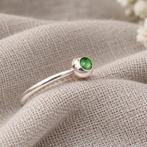 May include: A polished silver ring featuring a single, round, vibrant green gemstone set within a smooth, spherical metal casing. The band is slender and metallic, showcasing a clean, reflective surface that highlights the bright green stone.