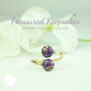 May include: A gold-toned bypass ring featuring two circular purple resin cabochons embedded with gold-colored flakes. The text "Treasured Keepsakes MEMORIAL JEWELLERY COLLECTION" is visible above the ring, along with a small bird icon.