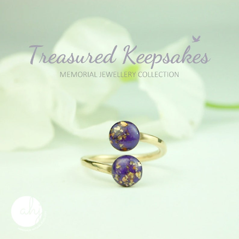 May include: A gold-toned bypass ring featuring two circular purple resin cabochons embedded with gold-colored flakes. The text "Treasured Keepsakes MEMORIAL JEWELLERY COLLECTION" is visible above the ring, along with a small bird icon.