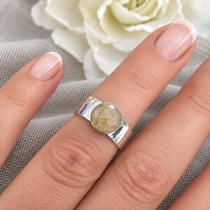 May include: A silver ring featuring a round, bezel-set rutilated quartz gemstone. The stone is translucent with visible golden needle-like inclusions throughout. The band is wide, smooth, and polished, with a reflective metallic finish.