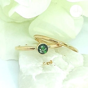 May include: Two gold rings. One ring features a small, round, green gemstone. The other ring is a simple gold band. The rings are displayed on a white surface with a blurred white floral background.