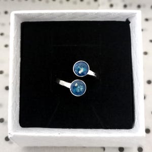 May include: A silver bypass ring featuring two circular settings, each containing a deep blue resin inlay with small white flecks. The ring is displayed inside a square white box with a black velvet insert, providing a clear view of the design.