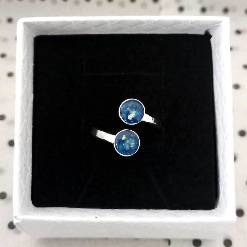 May include: A silver bypass ring featuring two circular settings, each containing a deep blue resin inlay with small white flecks. The ring is displayed inside a square white box with a black velvet insert, providing a clear view of the design.