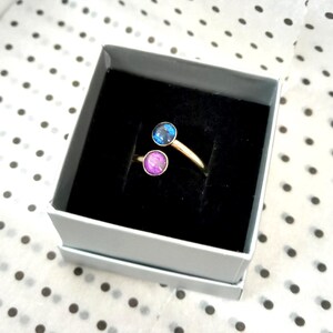 May include: A gold-toned open ring featuring two circular settings, one containing a dark blue iridescent stone and the other a vibrant purple iridescent stone, presented inside a square gray box with a black velvet interior lining.