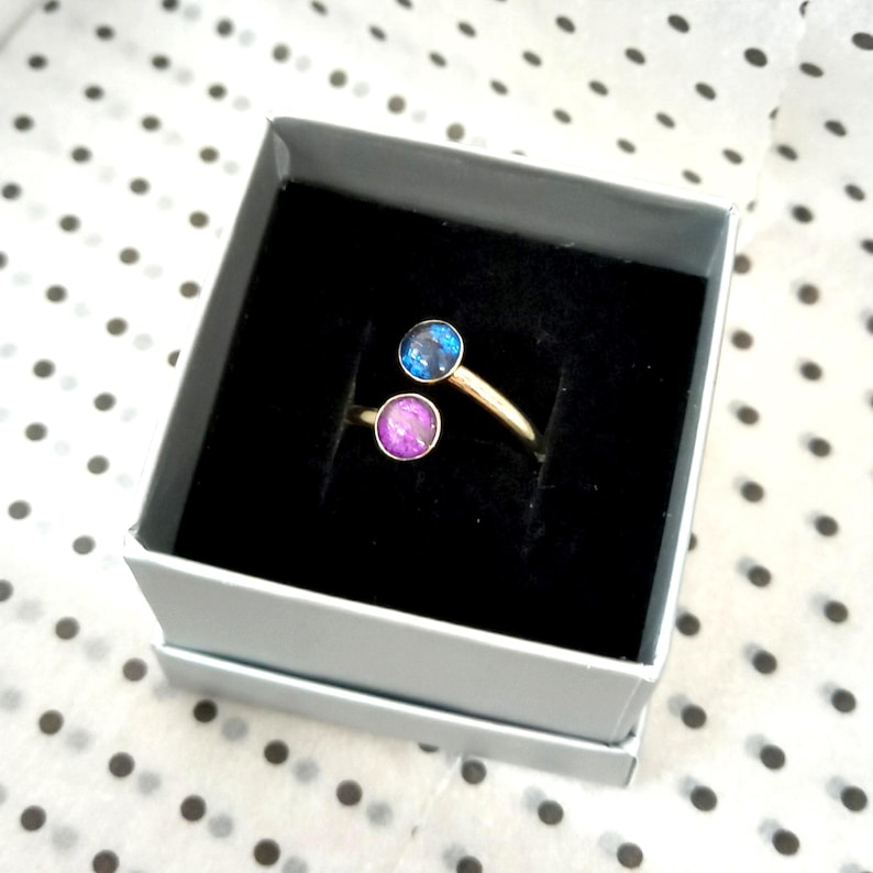 May include: A gold-toned open ring featuring two circular settings, one containing a dark blue iridescent stone and the other a vibrant purple iridescent stone, presented inside a square gray box with a black velvet interior lining.